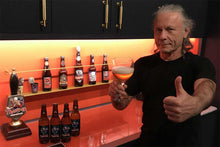 Chargez l&#39;image dans la visionneuse de la galerie, Bruce Dickinson giving a thumbs up while drinking Iron Maiden Trooper Basque Ale, with a display of Trooper beer bottles in the background.
