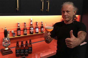 Bruce Dickinson giving a thumbs up while drinking Iron Maiden Trooper Basque Ale, with a display of Trooper beer bottles in the background.